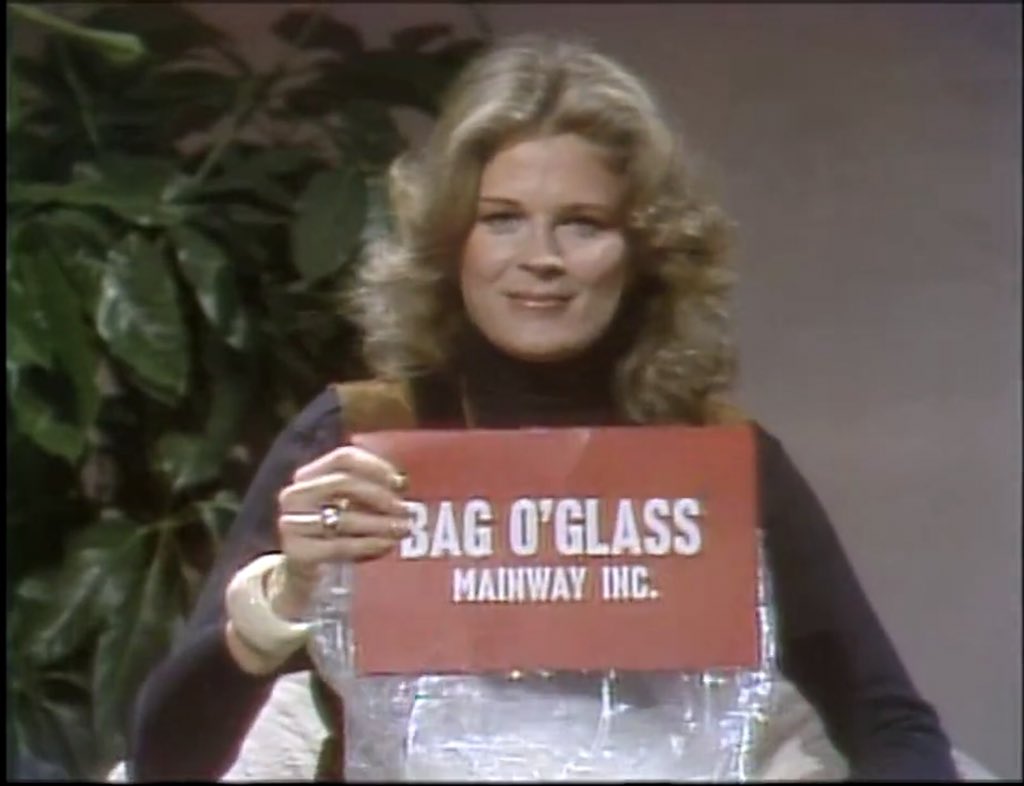 Woman holding a sign with 'Bag O'Glass Mainway Inc.' text against a plant and gray wall background.