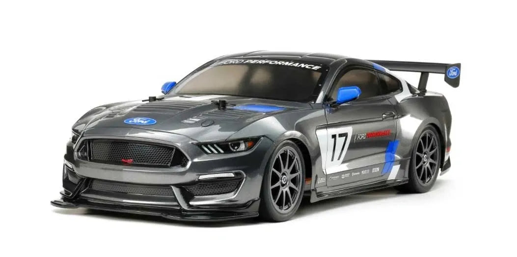 A Tamiya 1/10 scale model kit of a Ford Mustang GT4 4WD race car. The model has a gray body with blue and black detailing, a rear wing, and slick tires.
